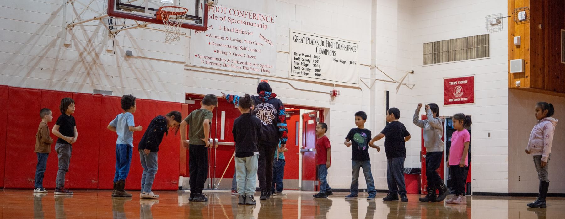 A large group of students standing together in a gymnasium, smiling and chatting, with basketball hoops in the background.