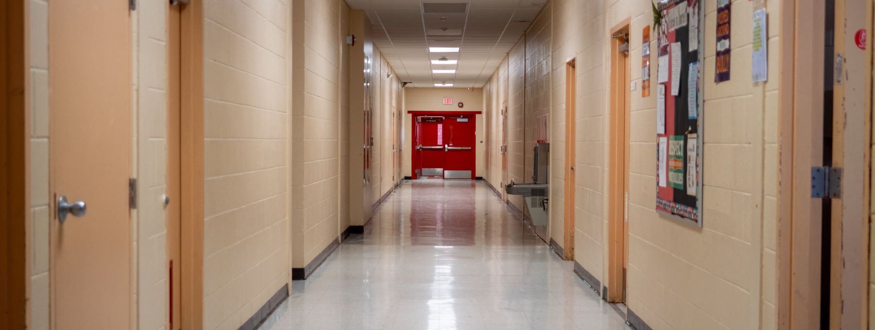 Looking down school hallway featuring a red exit door and lots of classroom doors flanking each side.